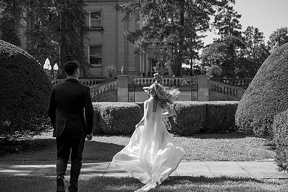 First look moment as bride in flowing wedding dress and veil taps groom’s shoulder on a stone terrace before a mansion and hedges