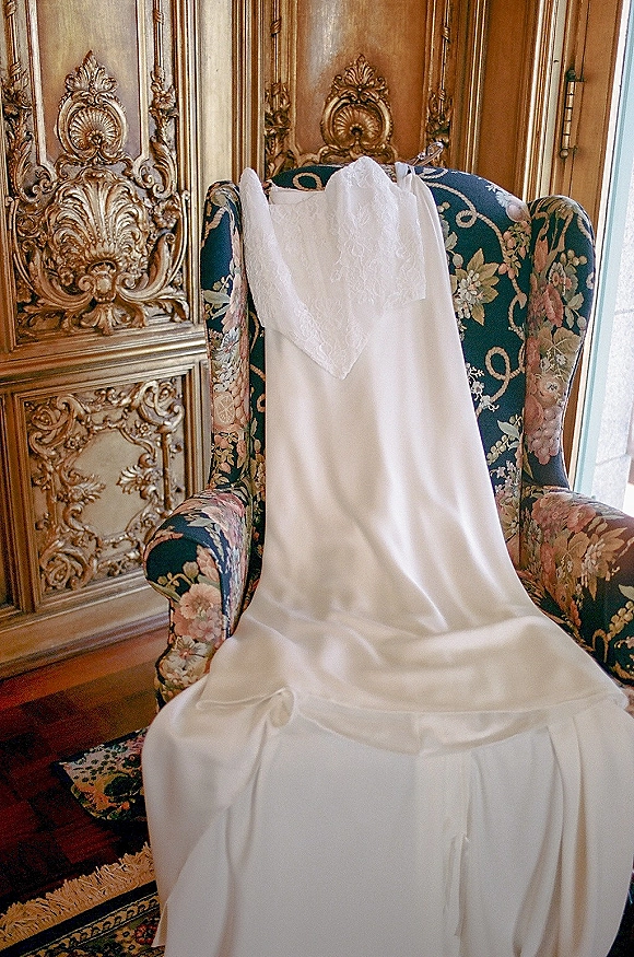 Wedding dress with lace wedding veil draped over a floral upholstered armchair, satin train spilling on a patterned rug in window light
