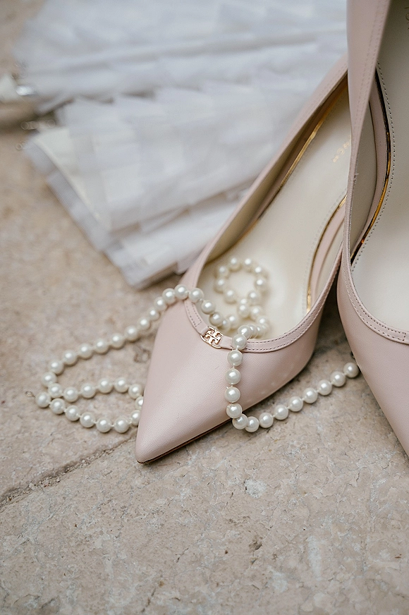Bridal shoes, blush wedding heels in pale pink with gold logo beside a pearl necklace and wedding invitation suite on stone surface