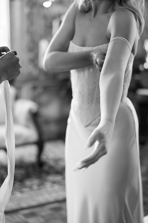 Bridal getting ready as bride putting on gloves beside her corset wedding dress, long gloves tied with a ribbon in window light