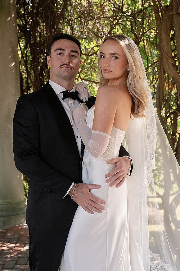 Couple portrait of bride and groom pose embracing in garden sunlight, bride in strapless dress with lace veil beside stone column