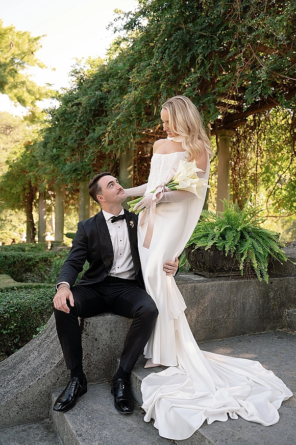 Couple portrait of bride in strapless gown with opera gloves holding white calla lily bouquet beside groom in black tux on stone steps by vine pergola
