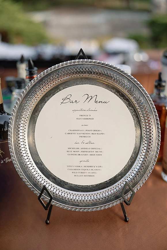 Wedding bar menu with signature cocktails sign displayed on a silver tray stand, calligraphy card beside liquor bottles at an outdoor bar table
