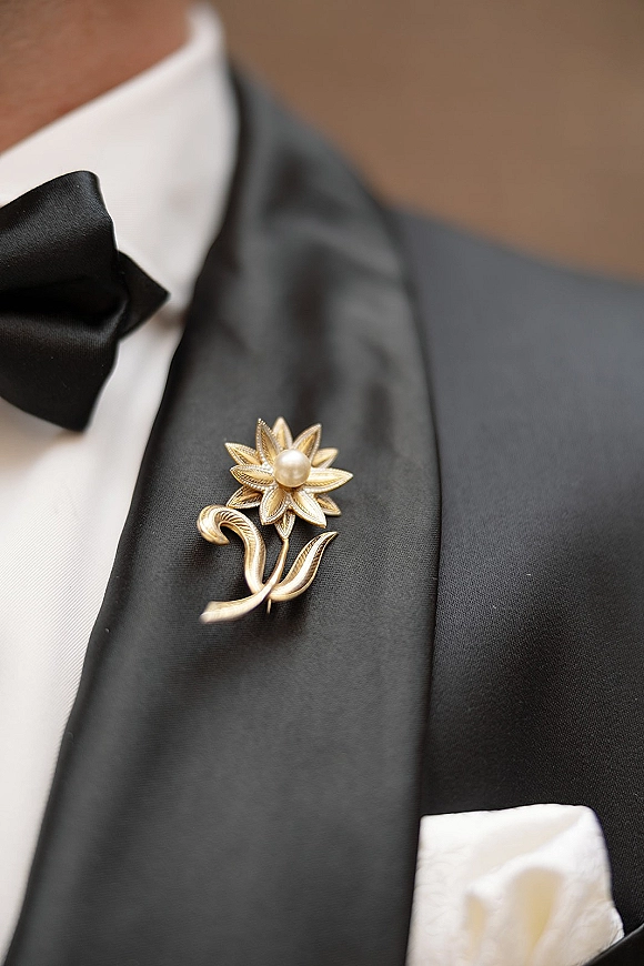 Groom boutonniere with gold lapel pin wedding detail, pearl-centered floral brooch on black satin tuxedo lapel with bow tie indoors