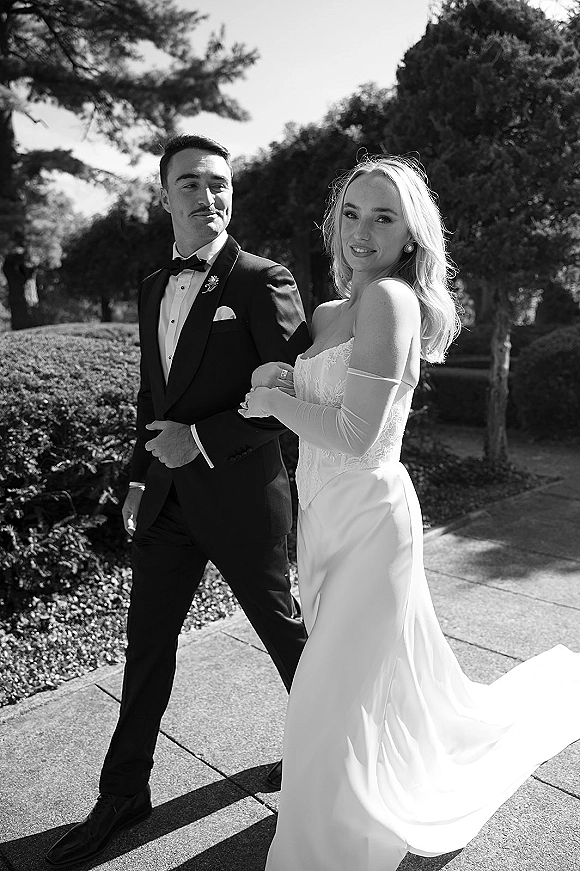 Couple portrait of bride and groom walking arm in arm, bride in lace bodice gown and long gloves, groom in tuxedo on a sunlit garden path
