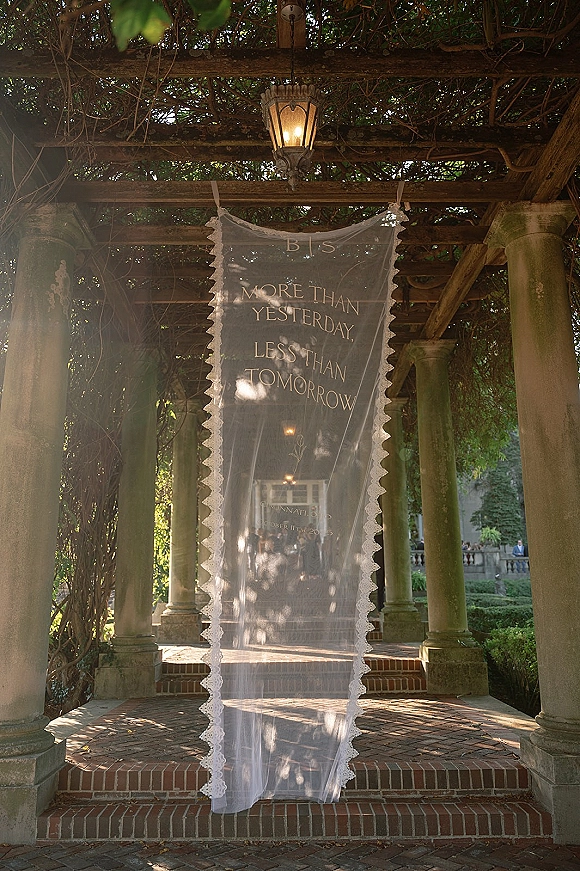 Wedding welcome sign with sheer fabric banner and calligraphy lettering, hanging by a lantern on a vine-covered garden pergola with stone columns