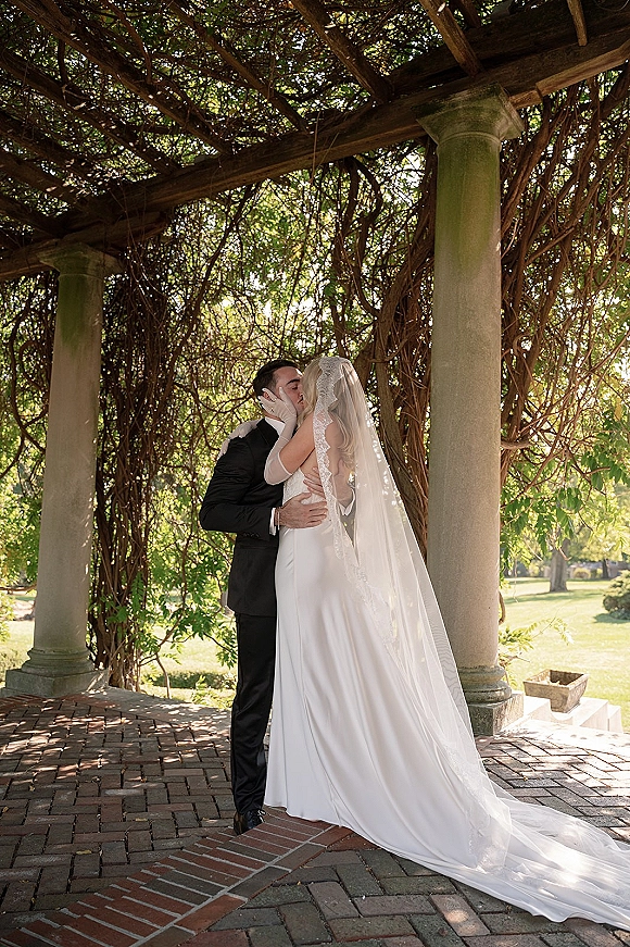 Wedding kiss as the bride and groom kiss under a vine-covered pergola, her lace-trim veil flowing behind his tuxedo on a brick patio