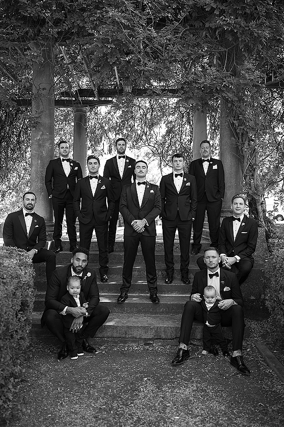 Groomsmen portrait with groom and groomsmen in black tuxedos and bow ties on stone steps under a vine-covered pergola with hedges
