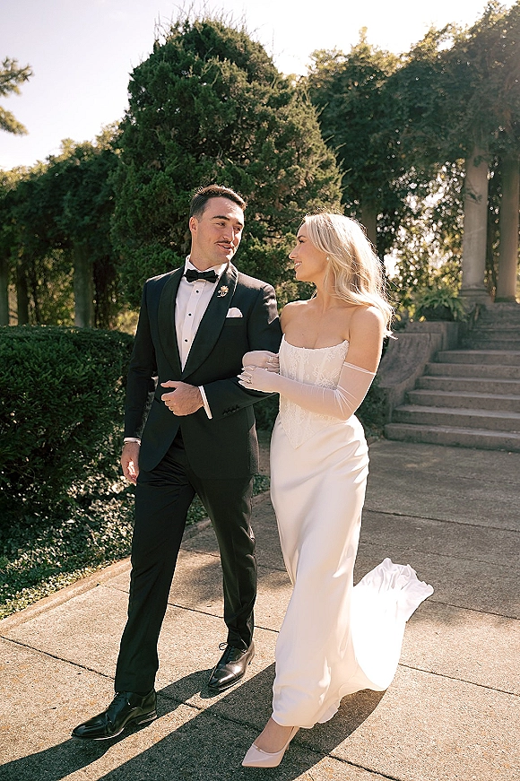Couple portrait of bride in a strapless gown with long gloves and groom in a black tuxedo, arm in arm on sunlit garden steps
