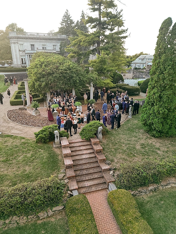 Wedding cocktail hour with outdoor cocktail hour tables draped in linens, greenery centerpieces and string lights on a formal garden terrace