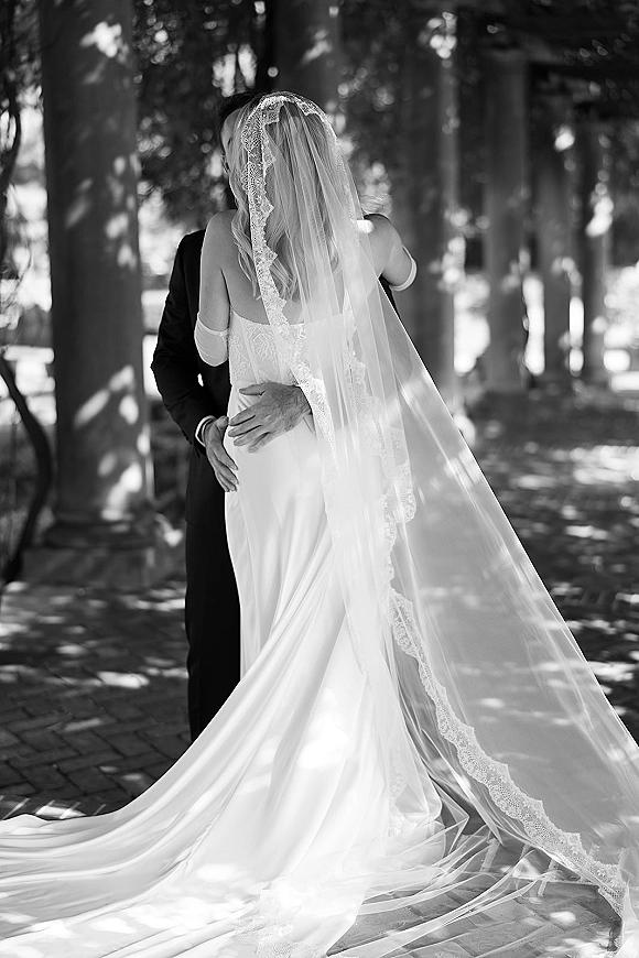 Wedding kiss as bride and groom kiss under a lace-trimmed veil on a shaded tree-lined walkway with dappled sunlight