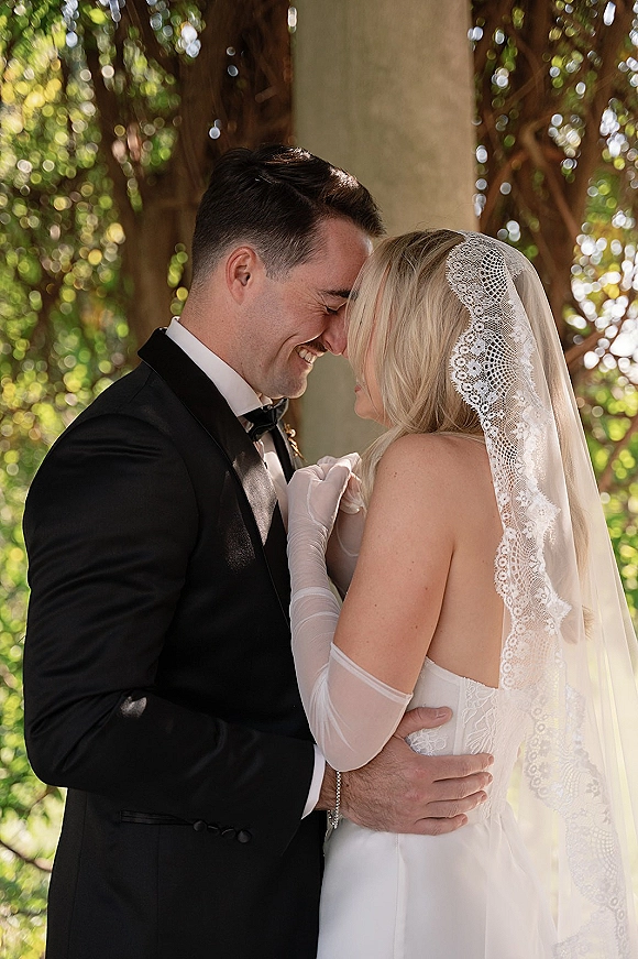 Couple portrait of bride and groom in a close embrace with a forehead touch, lace veil and tuxedo, framed by sunlit garden greenery
