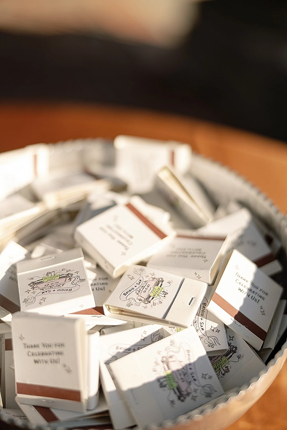 Wedding matchbooks in a bowl display with custom wedding matchbooks and printed packaging on a wood tabletop, blurred seating behind