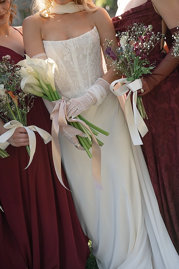 Bride with bridesmaids holding bouquets, bride in strapless lace wedding dress with calla lilies and ribbons in sunlit greenery outdoors