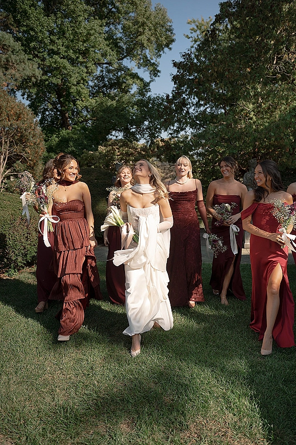 Bride with bridesmaids walking together, holding baby’s breath bouquets with white ribbon wrap on a garden lawn by stone steps under blue sky