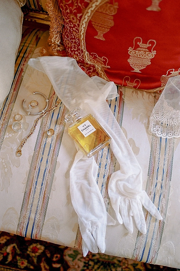 Bridal accessories flatlay with wedding day details: white opera gloves, perfume, gold bracelets and rings on lace atop a striped chair