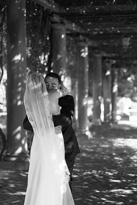 Wedding hug as bride and groom embrace, her lace-trimmed veil draped over his black suit on a pergola column walkway with trees