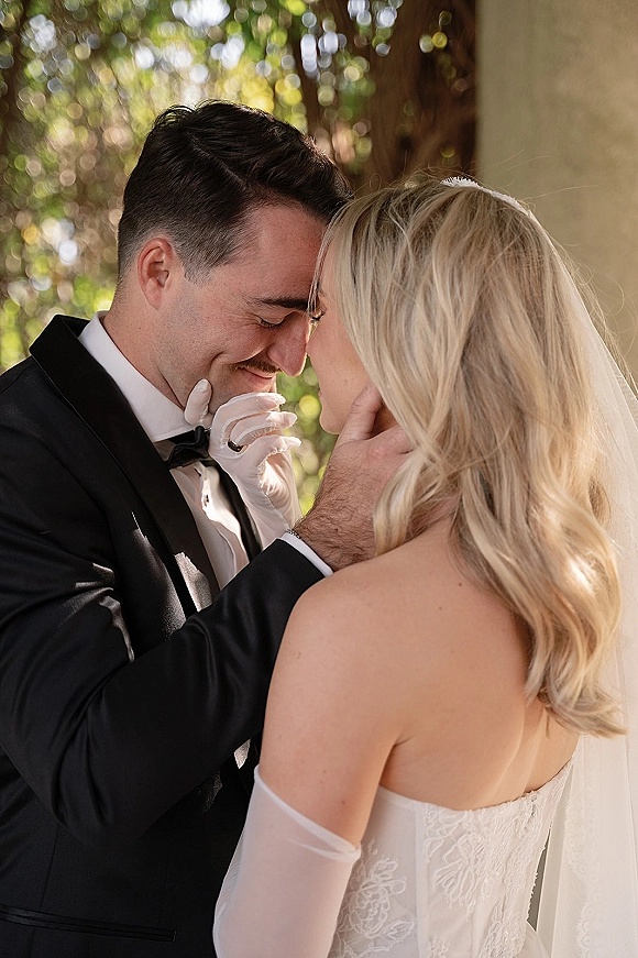 Wedding couple portrait with bride and groom forehead touch as he cradles her face, her lace dress, veil, and sheer gloves in sunlit trees backdrop
