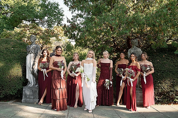 Bridesmaid group photo with bride and bridesmaids in burgundy bridesmaid dresses holding bouquets on a garden walkway by stone statues
