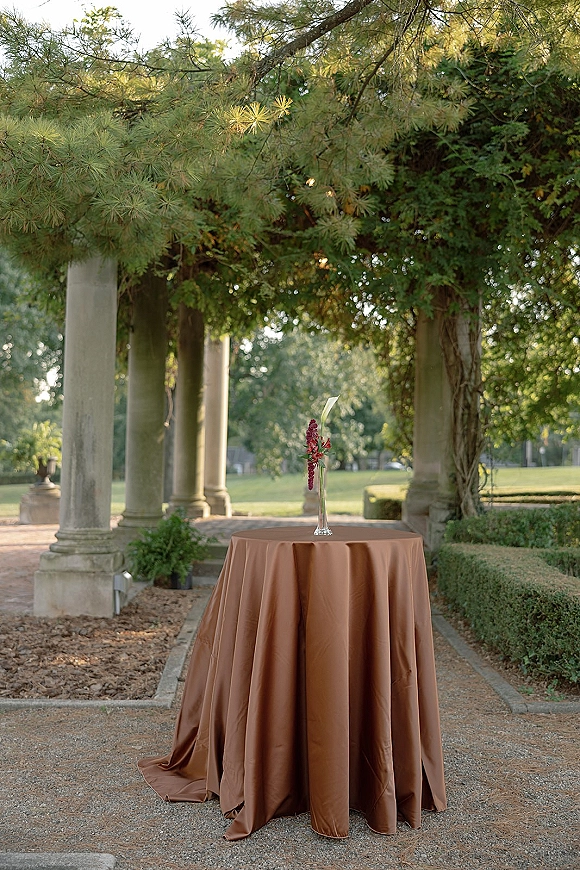 Cocktail table decor with brown tablecloth wedding linen, bud vase and single red floral stem near stone column pergola outdoors