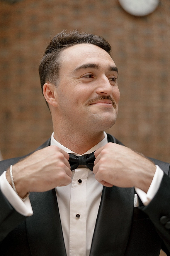 Groom portrait adjusting bow tie in a black tuxedo with shirt studs and pocket square, standing against a brick wall backdrop