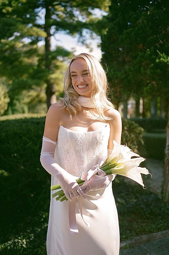 Bridal portrait of a smiling bride in a strapless lace wedding dress with long gloves, holding calla lilies in a sunlit garden