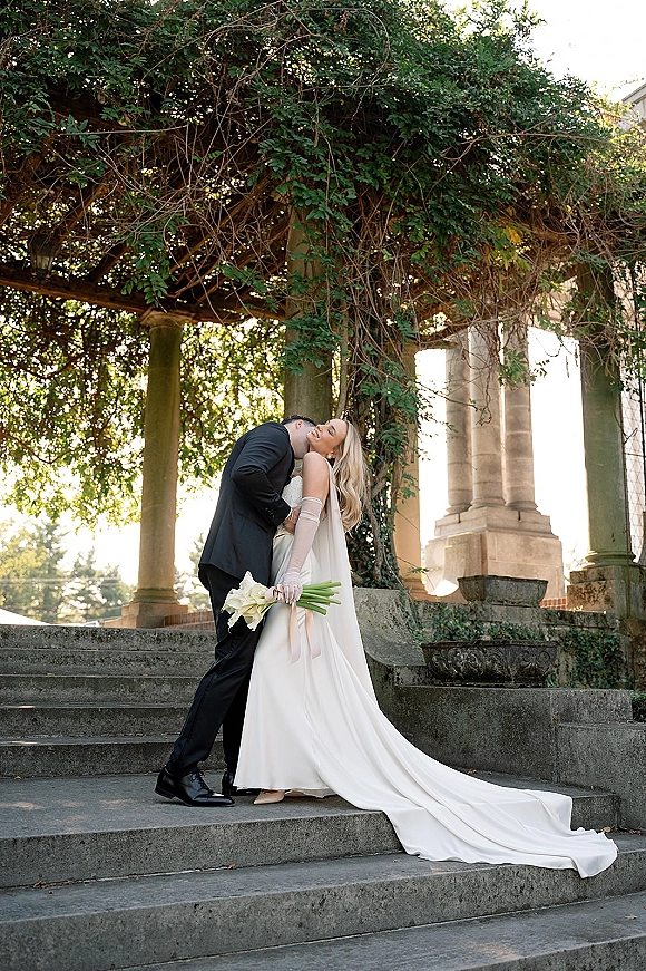 Wedding couple portrait of groom kissing bride’s cheek as she leans back holding calla lilies on stone steps by vine-covered columns in garden light