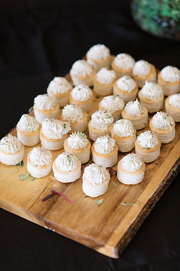 Wedding dessert bites arranged as mini cakes with whipped cream topping and herb garnish on a wooden serving board over a dark table surface