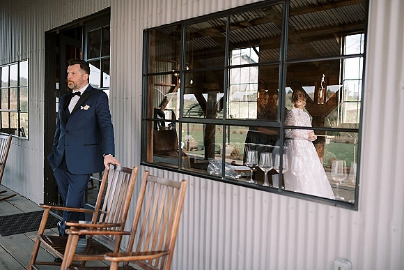 Wedding first look with groom waiting outside in a navy tuxedo as the bride stands behind black-framed window panes, separated by glass reflection