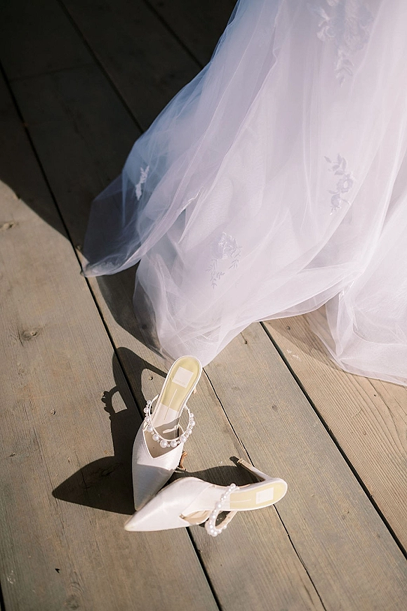 Bridal shoes, pearl wedding heels with pearl ankle straps and white pointed toes beside a tulle embroidered dress on a sunlit wood deck