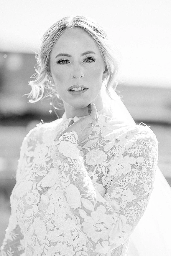 Bridal portrait in black and white of a bride with veil, hand on chin, wearing a high-neck long-sleeve lace gown and drop earrings outdoors
