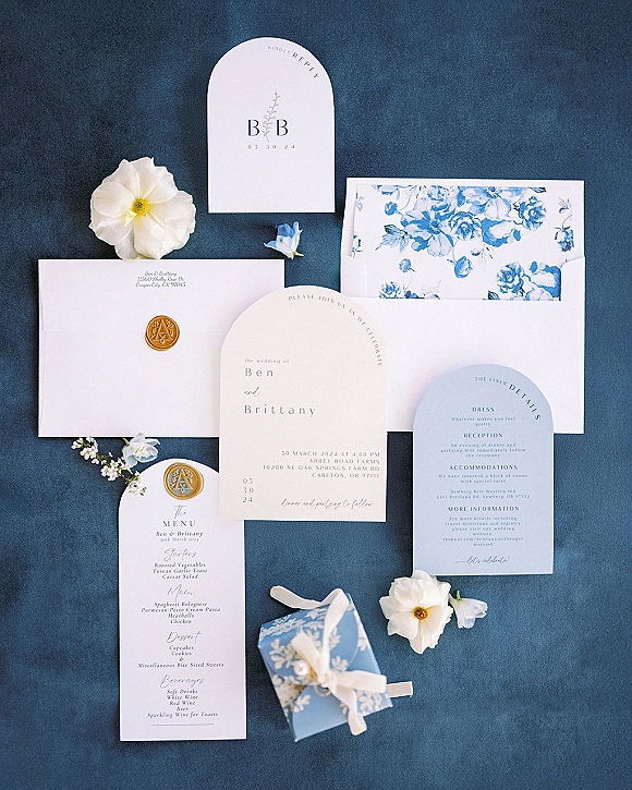Invitation suite wedding invitation flat lay with blue and white cards, gold wax seal, floral-lined envelopes, silk ribbon on blue fabric backdrop