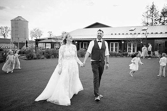 Wedding couple walking hand in hand, bride laughing in long sleeve lace dress and veil, groom in vest and bow tie on a barn lawn