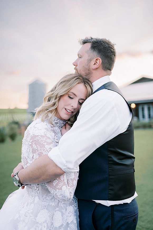 Couple portrait of bride and groom hug as he kisses her forehead in a field at sunset, with a barn and silo behind them