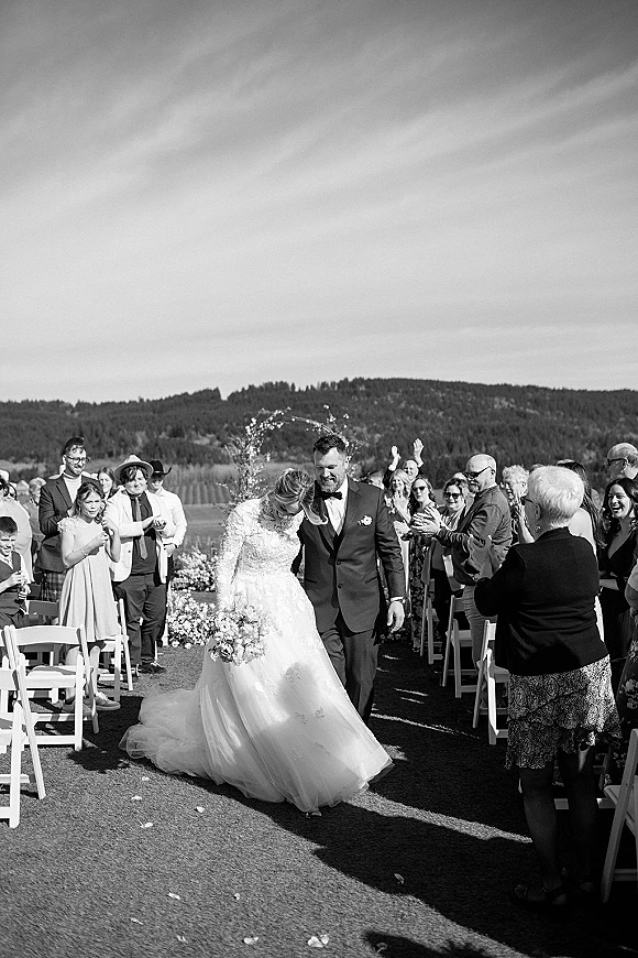 Wedding recessional as newlyweds walk down the aisle, bride in long sleeve lace gown holding bouquet, groom in black tux, guests cheering under floral arch