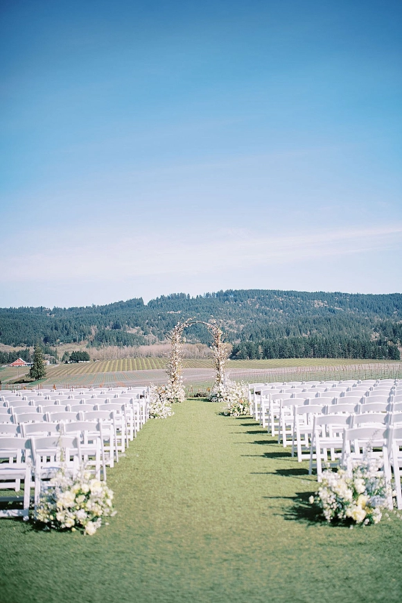 Ceremony setup for an outdoor wedding ceremony with a floral arch and white chairs lining a grassy aisle in a vineyard under blue sky