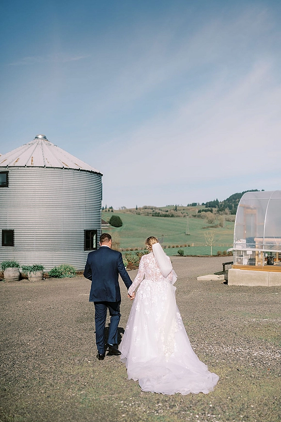 Couple portrait of newlyweds walking away hand in hand, bride in lace gown and long veil beside a grain silo on a gravel path