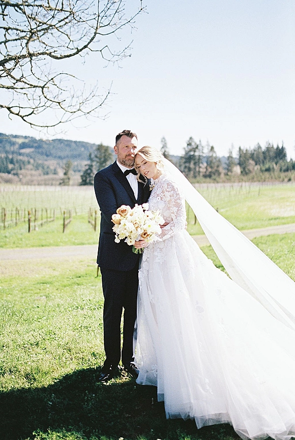 Couple portrait of bride and groom embrace, bride in lace dress with long veil holding bouquet in a vineyard with mountains behind