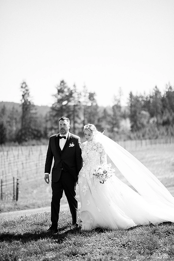 Couple portrait in a grassy field, bride in lace long sleeve wedding dress holding bouquet beside groom in tuxedo by fence line and hills