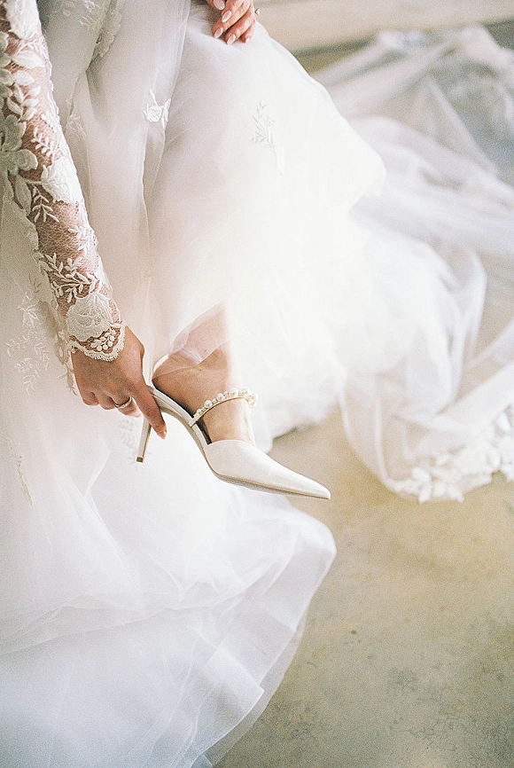 Bridal shoes in white wedding heels with pearl ankle straps as the bride adjusts them, lace sleeves and ring in soft interior light