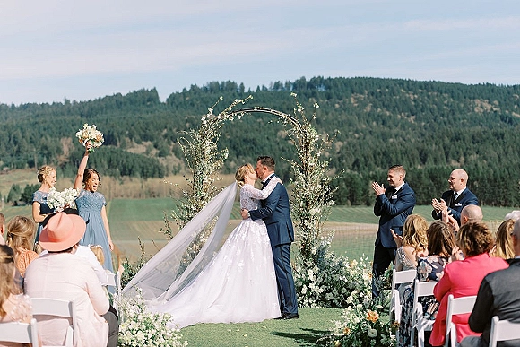 Ceremony kiss at an outdoor wedding ceremony as the bride and groom embrace under a floral arch with mountains behind them