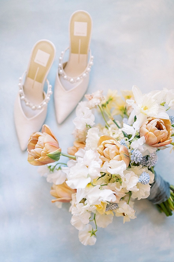 Bridal details flatlay with pearl wedding heels beside a peach tulip and white bouquet with blue accents on a light blue surface