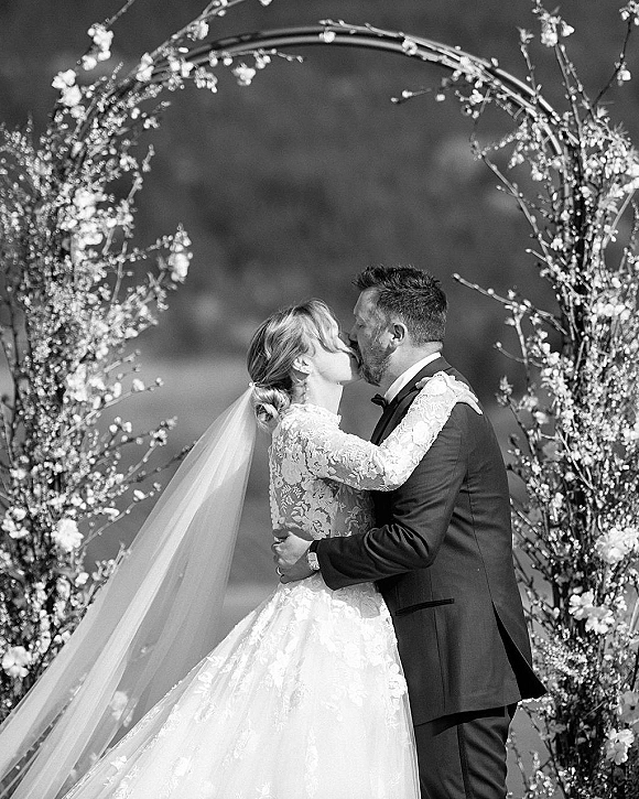 Wedding kiss portrait of bride and groom kissing beneath a floral arch, her lace dress and veil flowing as trees frame the outdoor scene