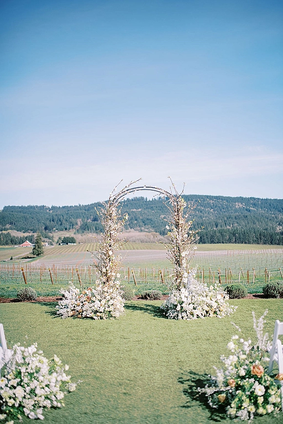 Ceremony arch decor with a circular wedding arch and white flowers, greenery, and aisle florals set on a vineyard lawn with hills
