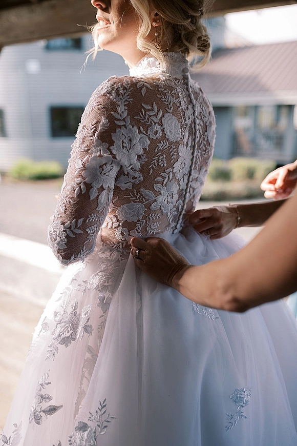 Wedding dress detail with lace wedding dress illusion sleeves and button back, tulle skirt and pearl earrings in daylight by windows