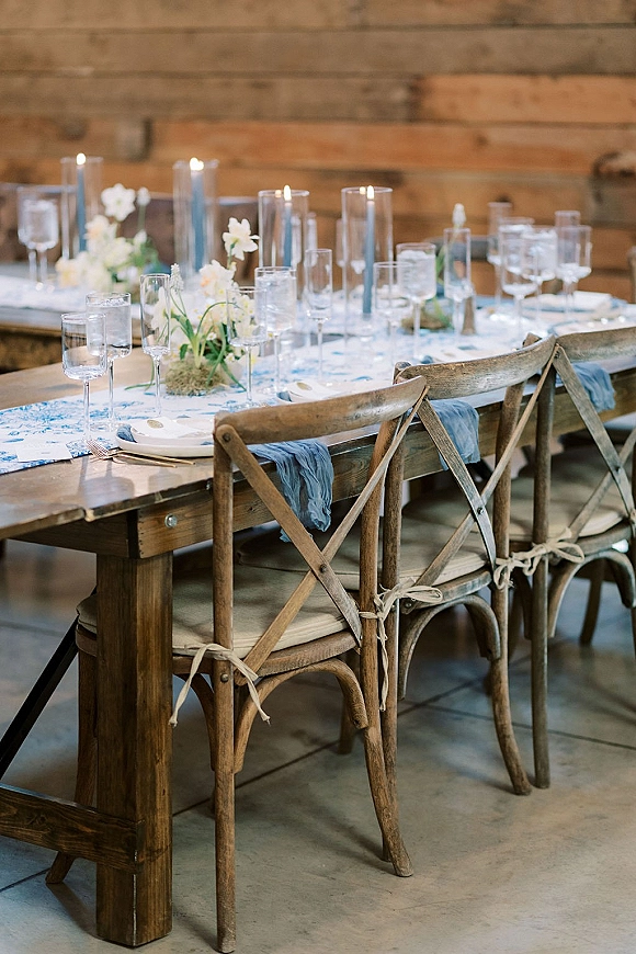 Reception tablescape with rustic wedding table setting on a wood farm table, blue napkins, white florals, and taper candles against a wood wall