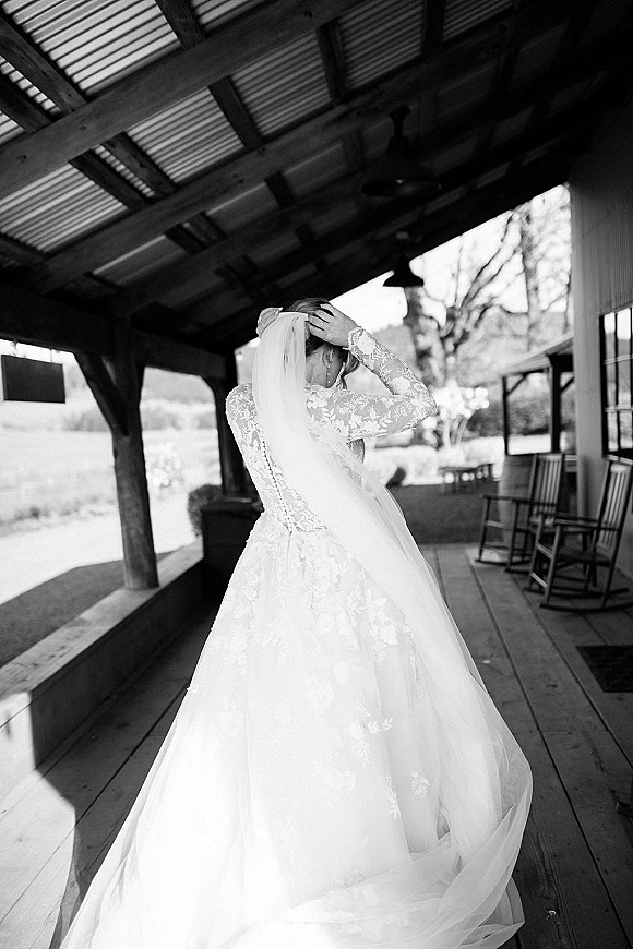 Bridal portrait of a bride adjusting her cathedral veil, showing a lace long sleeve wedding dress on a rustic covered porch with beams
