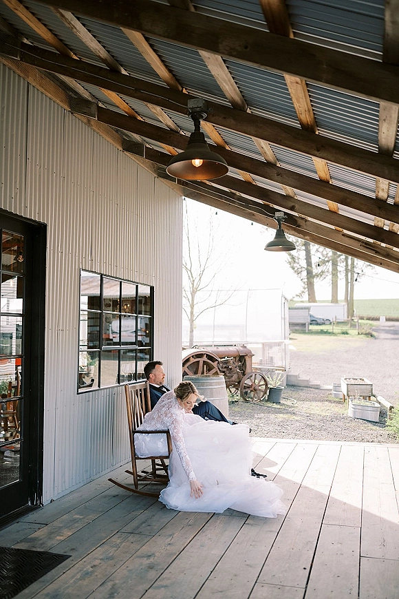 Couple portrait of bride and groom sitting on a rustic barn porch, her long-sleeve lace gown and tulle skirt beside him in a navy suit