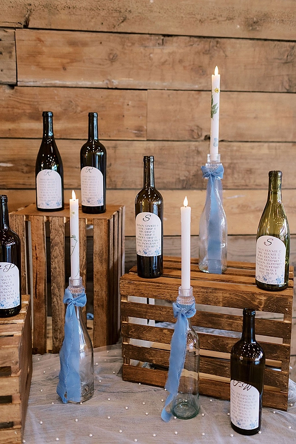 Wine bottle decor centerpiece with taper candles in bottles, blue ribbons, and labels arranged in wooden crates on a table by a wood plank wall