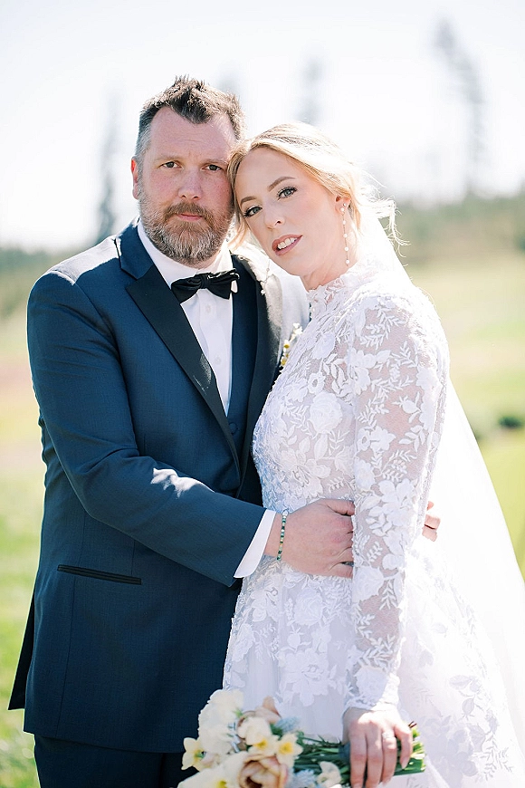 Couple portrait of bride and groom embrace, bride in lace long sleeve dress with veil and bouquet, groom in tuxedo in sunny field